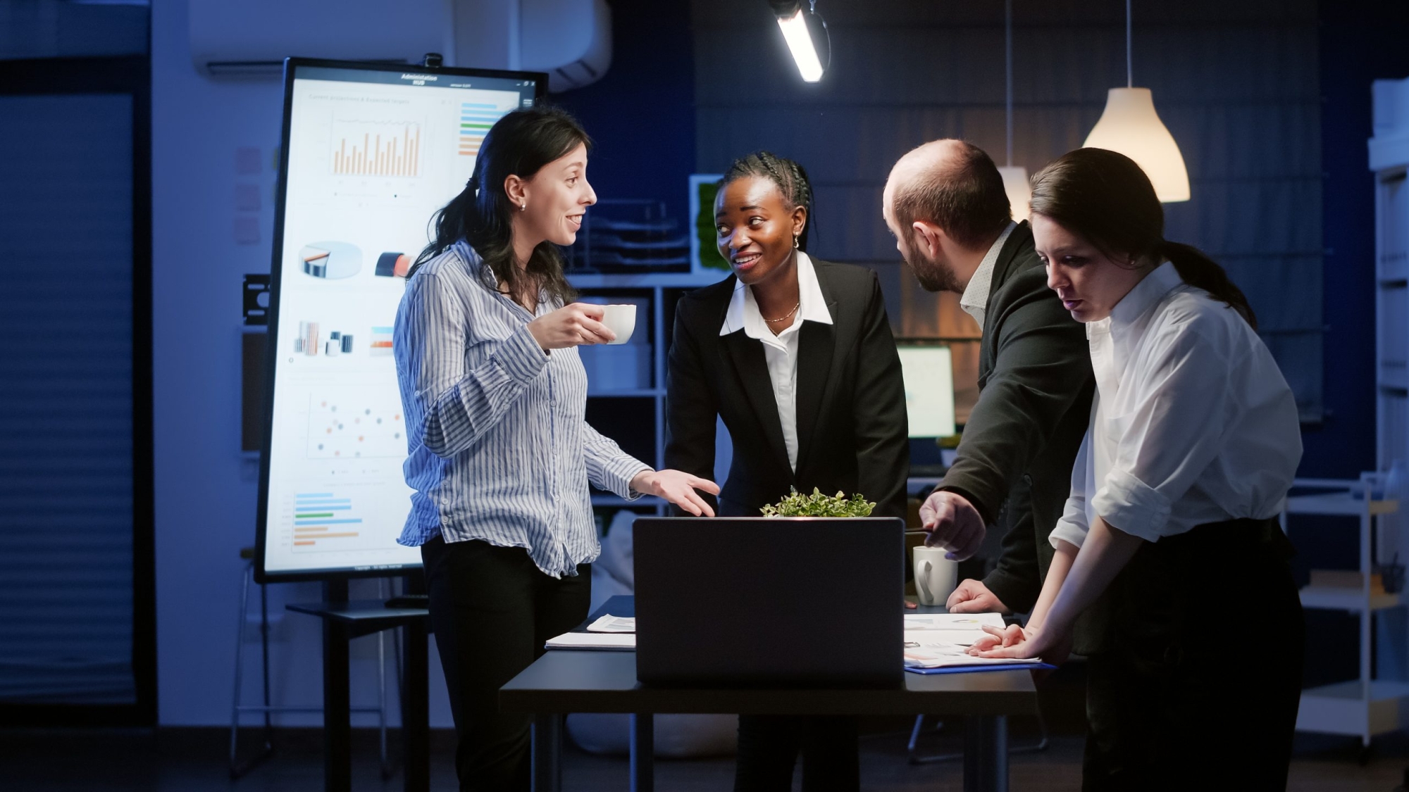 Excited diverse business teamwork receiving good news clapping while standing at table conference working in office meeting room late at night. Multi ethnic coworkers planning at company presentation